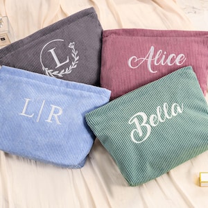 Peut inclure: Plusieurs trousses de maquillage en velours c&ocirc;tel&eacute; de couleurs vari&eacute;es : gris, rose, bleu et vert. Chaque trousse est brod&eacute;e d'un nom ou d'initiales en &eacute;criture blanche. Les trousses sont pos&eacute;es sur une surface cr&egrave;me.