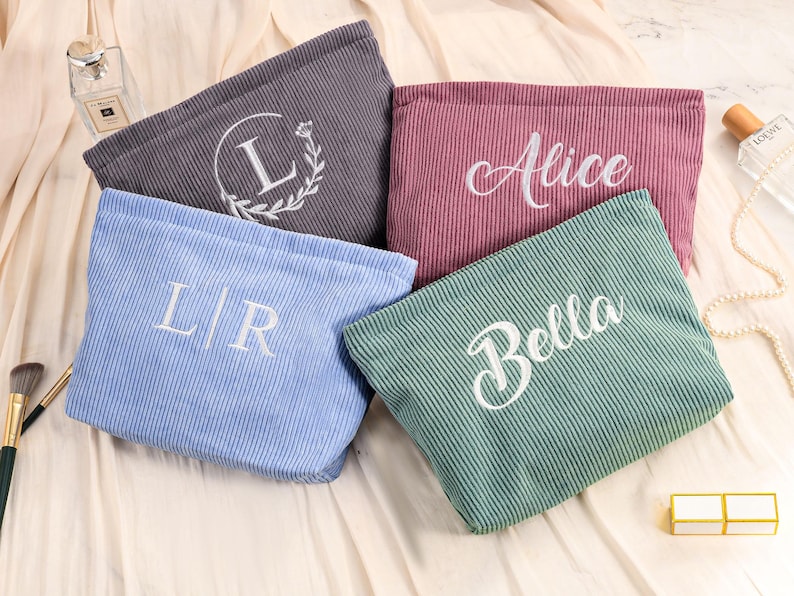 Peut inclure: Plusieurs trousses de maquillage en velours c&ocirc;tel&eacute; de couleurs vari&eacute;es : gris, rose, bleu et vert. Chaque trousse est brod&eacute;e d'un nom ou d'initiales en &eacute;criture blanche. Les trousses sont pos&eacute;es sur une surface cr&egrave;me.
