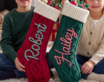 Op de afbeelding: Twee kerstsokken van kabelgebreid materiaal, één rood en één groen, elk met een witte geribbelde boord. De rode sok heeft de naam Robert in groen-witte touwletters, de groene sok heeft de naam Hailey in rood-witte touwletters.
