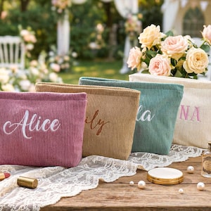 Peut inclure: Quatre trousses de maquillage en velours c&ocirc;tel&eacute; rose, beige, sarcelle et cr&egrave;me, chacune brod&eacute;e d'un nom dans une couleur compl&eacute;mentaire. Les sacs sont pr&eacute;sent&eacute;s sur une table en bois avec du maquillage et un flacon de parfum.