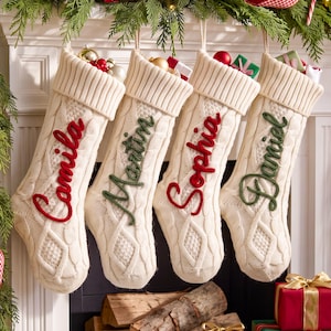 Puede incluir: Cuatro calcetines navideños de punto trenzado color crema colgados en fila. Cada calcetín tiene un puño acanalado y un nombre bordado en letra cursiva: Camila y Sophia en rojo, Martin y Daniel en verde. Pequeños regalos asoman por la parte superior.