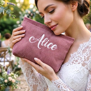 Peut inclure: Une pochette en velours c&ocirc;tel&eacute; rose poudr&eacute; avec le nom "Alice" brod&eacute; en &eacute;criture blanche. La pochette est tenue par une personne portant une robe en dentelle blanche, probablement lors d'un mariage. Le fond comprend des fleurs et des si&egrave;ges ext&eacute;rieurs.