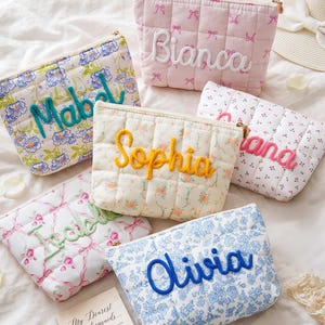 Könnte beinhalten: Sechs gesteppte Stofftaschen mit Reißverschluss, jede mit einem einzigartigen Blumen- oder Schleifenmuster und einem in erhabenem Garn gestickten Namen. Die Namen sind Mabel, Bianca, Sophia, Juliana, Isabelle und Olivia, dazu eine Karte mit My Dearest bridesmaids.