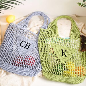 Puede incluir: Dos bolsas de malla de crochet, una azul claro y otra verde, con asas. La bolsa azul tiene las iniciales "CB" bordadas, y la bolsa verde tiene la inicial "K" bordada.