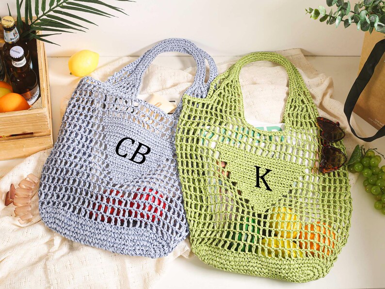 Puede incluir: Dos bolsas de malla de crochet, una azul claro y otra verde, con asas. La bolsa azul tiene las iniciales "CB" bordadas, y la bolsa verde tiene la inicial "K" bordada.
