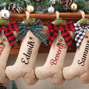 May include: Four burlap Christmas stockings with personalized names: Amanda, Edwin, Beverly, and Sammy. Each stocking has a plaid bow, greenery, and small bells. The stockings are decorated with red, green, and black plaid patterns.