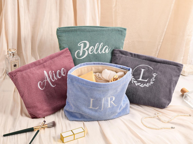Peut inclure: Quatre trousses de maquillage en velours c&ocirc;tel&eacute; rose, vert, bleu et gris. Les sacs ont du texte brod&eacute; blanc, comprenant des noms et des initiales. Un sac est ouvert, r&eacute;v&eacute;lant son contenu. Des pinceaux de maquillage et un rouge &agrave; l&egrave;vres sont au premier plan.