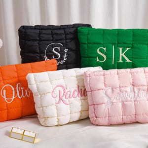 Peut inclure: Cinq trousses de maquillage matelass&eacute;es de couleurs vari&eacute;es : noir, vert, orange, blanc et rose. Chaque sac comporte du texte ou des initiales brod&eacute;s dans une couleur contrastante. Les sacs sont rectangulaires avec un design rembourr&eacute; et cousu.