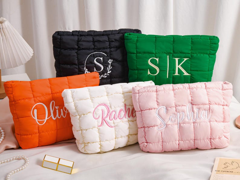 Peut inclure: Cinq trousses de maquillage matelass&eacute;es de couleurs vari&eacute;es : noir, vert, orange, blanc et rose. Chaque sac comporte du texte ou des initiales brod&eacute;s dans une couleur contrastante. Les sacs sont rectangulaires avec un design rembourr&eacute; et cousu.