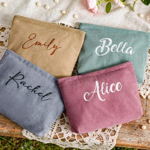 Peut inclure: Quatre pochettes en velours c&ocirc;tel&eacute; de couleurs beige, turquoise, rose poudr&eacute; et grise, chacune brod&eacute;e d'un pr&eacute;nom diff&eacute;rent : "Emily", "Bella", "Alice" et "Rachel". Les pochettes sont dispos&eacute;es sur une surface en bois rustique avec des dentelles et des perles.