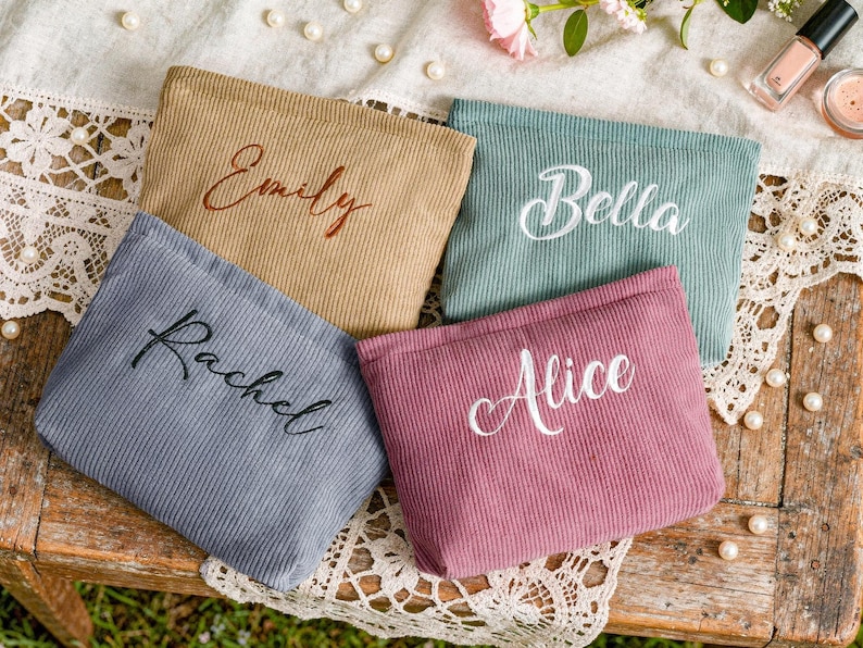Peut inclure: Quatre pochettes en velours c&ocirc;tel&eacute; de couleurs beige, turquoise, rose poudr&eacute; et grise, chacune brod&eacute;e d'un pr&eacute;nom diff&eacute;rent : "Emily", "Bella", "Alice" et "Rachel". Les pochettes sont dispos&eacute;es sur une surface en bois rustique avec des dentelles et des perles.