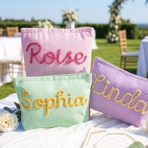 May include: Three seersucker cosmetic bags in pink, green, and purple, each with a name embroidered in a contrasting color. The bags are on a white lace-covered table, with a champagne bottle and glasses in the background. The names embroidered are "Rose", "Sophia", and "Linda".