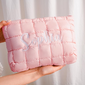 Peut inclure: Une pochette matelass&eacute;e rose clair avec le nom "Sophia" brod&eacute; en &eacute;criture blanche. La pochette a une forme rectangulaire et une texture gonfl&eacute;e et rembourr&eacute;e. Le fond est de couleur neutre.