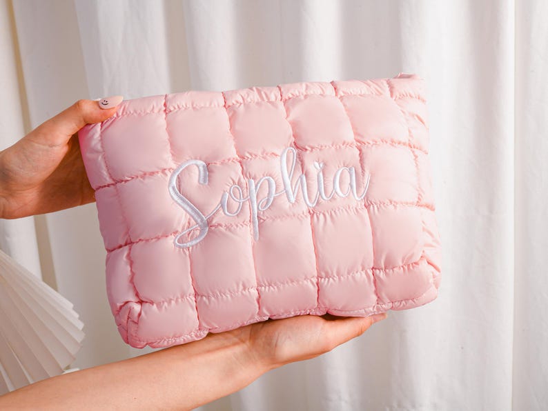 Peut inclure: Une pochette matelass&eacute;e rose clair avec le nom "Sophia" brod&eacute; en &eacute;criture blanche. La pochette a une forme rectangulaire et une texture gonfl&eacute;e et rembourr&eacute;e. Le fond est de couleur neutre.