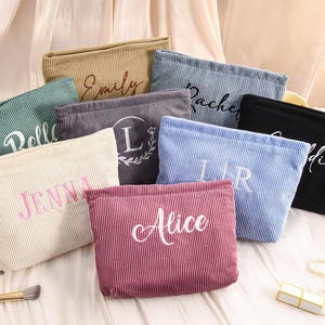 Peut inclure: Plusieurs trousses de maquillage en velours c&ocirc;tel&eacute; de diff&eacute;rentes couleurs, notamment beige, vert, gris, bleu et noir. Chaque trousse est brod&eacute;e d'un nom ou d'une initiale de couleur contrastante. Les trousses sont dispos&eacute;es sur une surface claire.