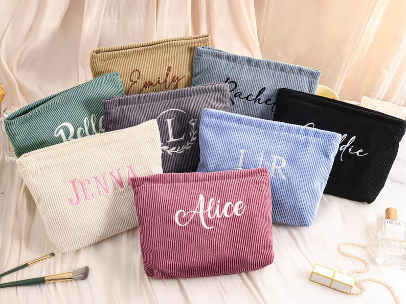 Peut inclure: Plusieurs trousses de maquillage en velours c&ocirc;tel&eacute; de diff&eacute;rentes couleurs, notamment beige, vert, gris, bleu et noir. Chaque trousse est brod&eacute;e d'un nom ou d'une initiale de couleur contrastante. Les trousses sont dispos&eacute;es sur une surface claire.