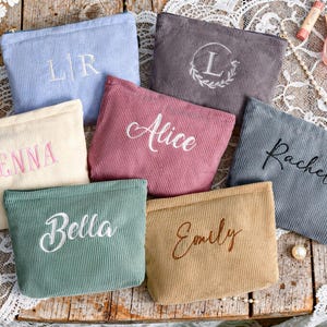 Peut inclure: Plusieurs trousses de maquillage en velours c&ocirc;tel&eacute; de diff&eacute;rentes couleurs, dont bleu, rose et beige. Chaque trousse pr&eacute;sente du texte brod&eacute;, comme des noms et des initiales. Les trousses sont dispos&eacute;es sur une surface en bois avec des produits de beaut&eacute; et des perles.