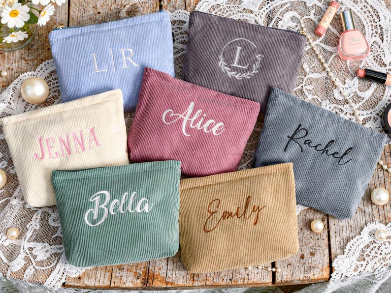 Peut inclure: Plusieurs trousses de maquillage en velours c&ocirc;tel&eacute; de diff&eacute;rentes couleurs, dont bleu, rose et beige. Chaque trousse pr&eacute;sente du texte brod&eacute;, comme des noms et des initiales. Les trousses sont dispos&eacute;es sur une surface en bois avec des produits de beaut&eacute; et des perles.