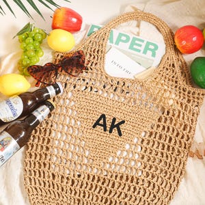 Puede incluir: Una bolsa de mano de crochet de color beige con las iniciales "AK" bordadas en la parte delantera. La bolsa est&aacute; sobre una superficie blanca con fruta y otros objetos alrededor.