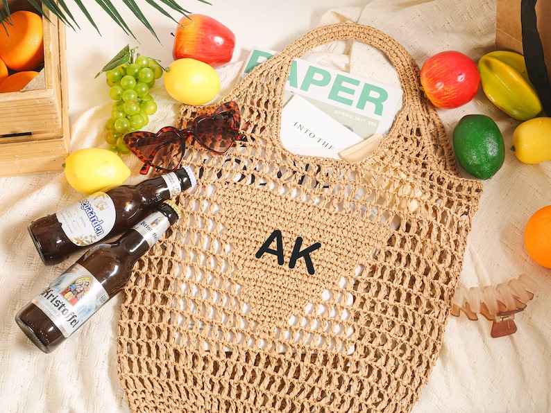 Puede incluir: Una bolsa de mano de crochet de color beige con las iniciales "AK" bordadas en la parte delantera. La bolsa est&aacute; sobre una superficie blanca con fruta y otros objetos alrededor.