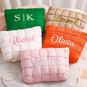 Peut inclure: Cinq coussins carr&eacute;s et matelass&eacute;s de diff&eacute;rentes couleurs : vert, beige, blanc, orange et rose. Chaque coussin porte une broderie avec des noms et des initiales. Les coussins sont dispos&eacute;s sur un lit avec d'autres objets d&eacute;coratifs.