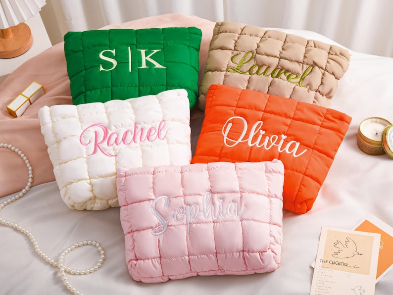 Peut inclure: Cinq coussins carr&eacute;s et matelass&eacute;s de diff&eacute;rentes couleurs : vert, beige, blanc, orange et rose. Chaque coussin porte une broderie avec des noms et des initiales. Les coussins sont dispos&eacute;s sur un lit avec d'autres objets d&eacute;coratifs.