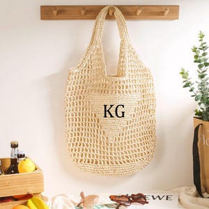 Puede incluir: Una bolsa de mano de crochet beige con un tejido grande y abierto. La bolsa tiene un bolsillo grande en la parte delantera con la palabra "KG" impresa en negro.