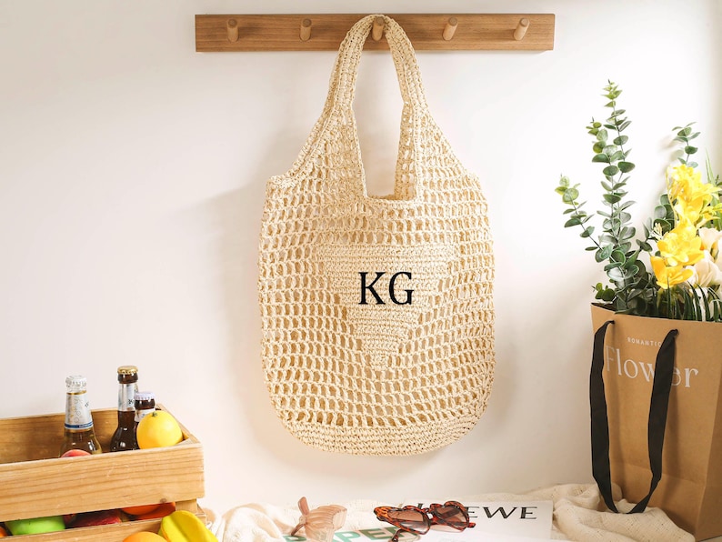 Puede incluir: Una bolsa de mano de crochet beige con un tejido grande y abierto. La bolsa tiene un bolsillo grande en la parte delantera con la palabra "KG" impresa en negro.
