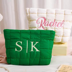 Peut inclure: Deux trousses de maquillage matelass&eacute;es. L'une est vert &eacute;meraude avec les initiales "S|K" brod&eacute;es en blanc. L'autre est blanc cass&eacute; avec le nom "Rachel" brod&eacute; en rose. Les deux sacs ont un motif carr&eacute;.