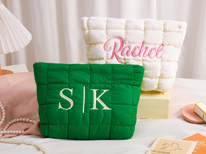 Peut inclure: Deux trousses de maquillage matelass&eacute;es. L'une est vert &eacute;meraude avec les initiales "S|K" brod&eacute;es en blanc. L'autre est blanc cass&eacute; avec le nom "Rachel" brod&eacute; en rose. Les deux sacs ont un motif carr&eacute;.