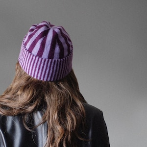 Puede incluir: Un gorro de punto a rayas moradas y lavanda. El gorro lo lleva una persona con pelo largo castaño y una chaqueta de cuero negra. El gorro tiene un borde acanalado y rayas verticales.