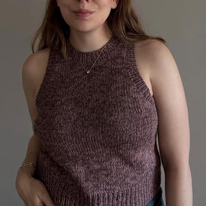 May include: A sleeveless, knitted top in a muted burgundy color. The top has a ribbed texture and a high neckline. A delicate silver necklace with a small pendant is visible. The background is a solid, neutral gray.