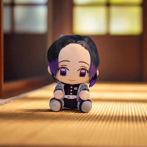 Op de afbeelding: Een pluche pop van een anime-personage met zwart haar en paarse highlights. De pop draagt een zwart-witte outfit met een paars-wit jack met patroon. De pop zit op een lichtbruin oppervlak.