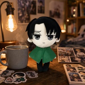 Puede incluir: Muñeco de peluche de un personaje con cabello negro, capa verde y pantalones negros. El muñeco está sobre una superficie de madera junto a una taza de café y pegatinas. El fondo incluye una lámpara y una estantería.