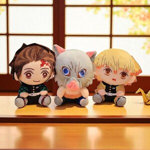 Op de afbeelding: Drie pluche poppen van anime-personages. E&eacute;n heeft bruin haar en een litteken, een andere een varkensachtig hoofd en de derde geel haar. Ze zitten op een houten oppervlak. De poppen hebben grote ogen en gedetailleerde gezichtskenmerken.