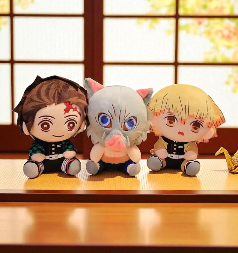 Op de afbeelding: Drie pluche poppen van anime-personages. E&eacute;n heeft bruin haar en een litteken, een andere een varkensachtig hoofd en de derde geel haar. Ze zitten op een houten oppervlak. De poppen hebben grote ogen en gedetailleerde gezichtskenmerken.