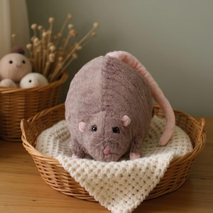 Può includere: Un giocattolo di peluche rotondo a forma di ratto con morbida pelliccia marrone polveroso, piccole orecchie rosa, naso rosa e una lunga coda rosa curva. L'animale di pezza è posizionato all'interno di un cesto di vimini intrecciato.