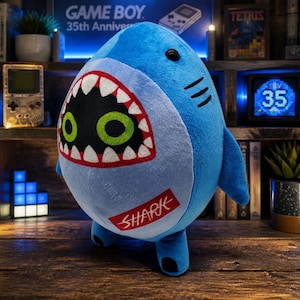 May include: A plush shark toy with a rounded blue body, a light blue belly, and a wide open mouth featuring white teeth and large green eyes. A red rectangular patch with the white text SHARK is stitched onto the lower belly area.