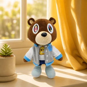 Puede incluir: Un peluche de oso marrón que viste una chaqueta universitaria gris y blanca con ribetes azules sobre una camiseta blanca con un gráfico amarillo. El oso tiene ojos grandes y estilizados con detalles coloridos, nariz negra y zapatos azul claro.