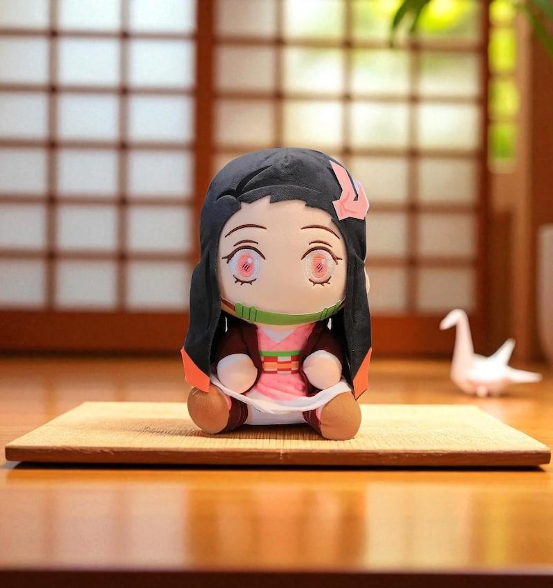 Op de afbeelding: Pluche pop van een anime-personage met zwart haar, roze ogen en een groene mondbeschermer. De pop draagt een roze kimono met een bruine jas en zit op een kleine mat. Een witte origami kraanvogel staat op de achtergrond.