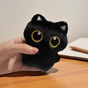Puede incluir: Un juguete de peluche negro con forma de gato con grandes ojos amarillos y detalles blancos en las orejas. El gato tiene una forma redondeada y está hecho de material suave y difuso. El juguete se sostiene en una mano.