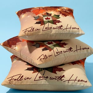 Puede incluir: Una pila de tres cojines cuadrados de color beige con un diseño estampado de hojas de otoño en tonos naranja, marrón y verde. Cada cojín muestra el texto "Fall in Love with Home" en una fuente cursiva de color marrón oscuro.