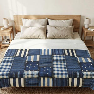 Op de afbeelding: Een patchwork sprei met een verscheidenheid aan blauwe en witte patronen, waaronder strepen, vichy-ruitjes en stippen. De sprei ligt over een bed met een wit laken, twee beige kussens en een houten hoofdeinde in een slaapkameromgeving.