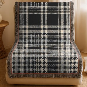 May include: A rectangular woven throw blanket featuring a black, white, and gray plaid pattern with a houndstooth design element. The blanket has a decorative fringe along the edges and is draped over a neutral-colored upholstered chair.