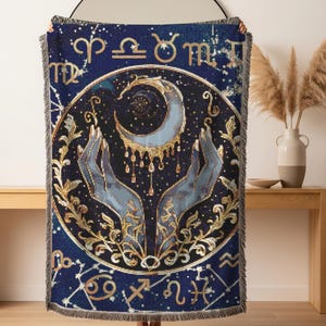 Puede incluir: Una manta tejida rectangular con fondo azul oscuro y motivos celestiales dorados. El centro muestra dos manos extendidas hacia una luna creciente con gotas doradas colgantes, rodeadas por un marco circular con símbolos del zodiaco.