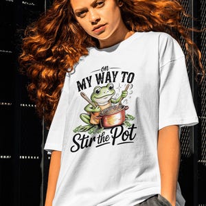 Puede incluir: Camiseta blanca con un diseño de rana de dibujos animados removiendo una olla. El texto "On My Way To Stir the Pot" está presente. La camiseta tiene un corte holgado y es adecuada para uso casual.