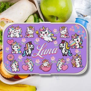 Puede incluir: Una fiambrera rectangular de color morado con un patrón repetitivo de unicornios de dibujos animados, pequeñas flores y el nombre Luna escrito en letra blanca. Los unicornios aparecen en varias poses, sosteniendo corazones y helados.