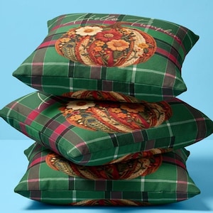 Puede incluir: Una pila de tres cojines cuadrados con un patrón de cuadros en verde, rojo y negro. Cada cojín muestra un gráfico central de una calabaza decorada con flores coloridas y el texto Hello Pumpkin en letra cursiva de color blanco.