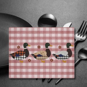 Puede incluir: Un mantel individual rectangular con un patrón de cuadros rojos y blancos. Se presentan tres patos decorativos con alas estampadas y cabezas verdes. Pequeños acentos de flores marrones están dispersos alrededor de los patos. Cubiertos visibles en el fondo.