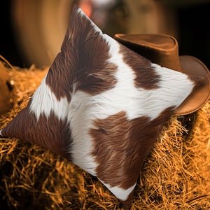 Puede incluir: Un cojín decorativo cuadrado con un diseño de piel de vaca en tonos marrón y blanco. Descansa sobre un montón de heno, con un sombrero de vaquero marrón en el fondo. El cojín tiene un aspecto suave y texturizado.
